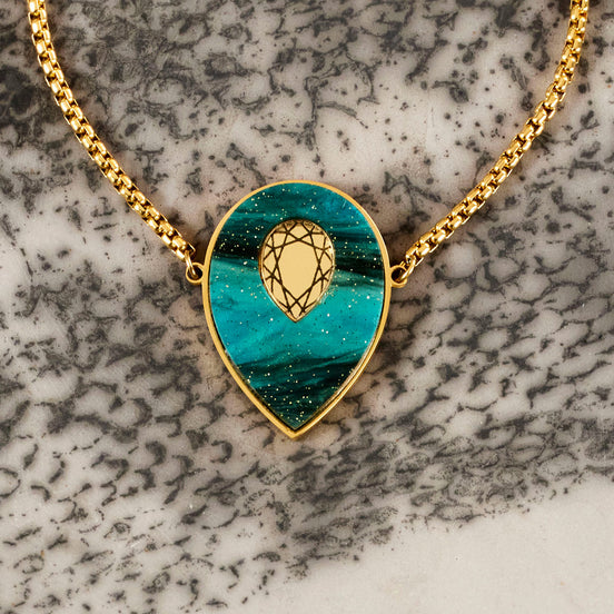 A gold-colored bolo chain bracelet with a teal marbled agate pendant and a mirrored gold gemstone in the center, displayed on a textured background.