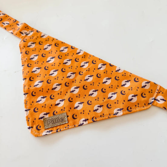 An orange triangular pet bandana with a repeating black and white crescent moon and star pattern, featuring a small brown leather brand patch that reads 'Pawme'.