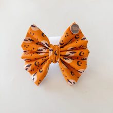 Load image into Gallery viewer, An orange fabric hair bow with a Halloween pattern of black bats, white moons, and small stars, displayed on a white stand against a light background.