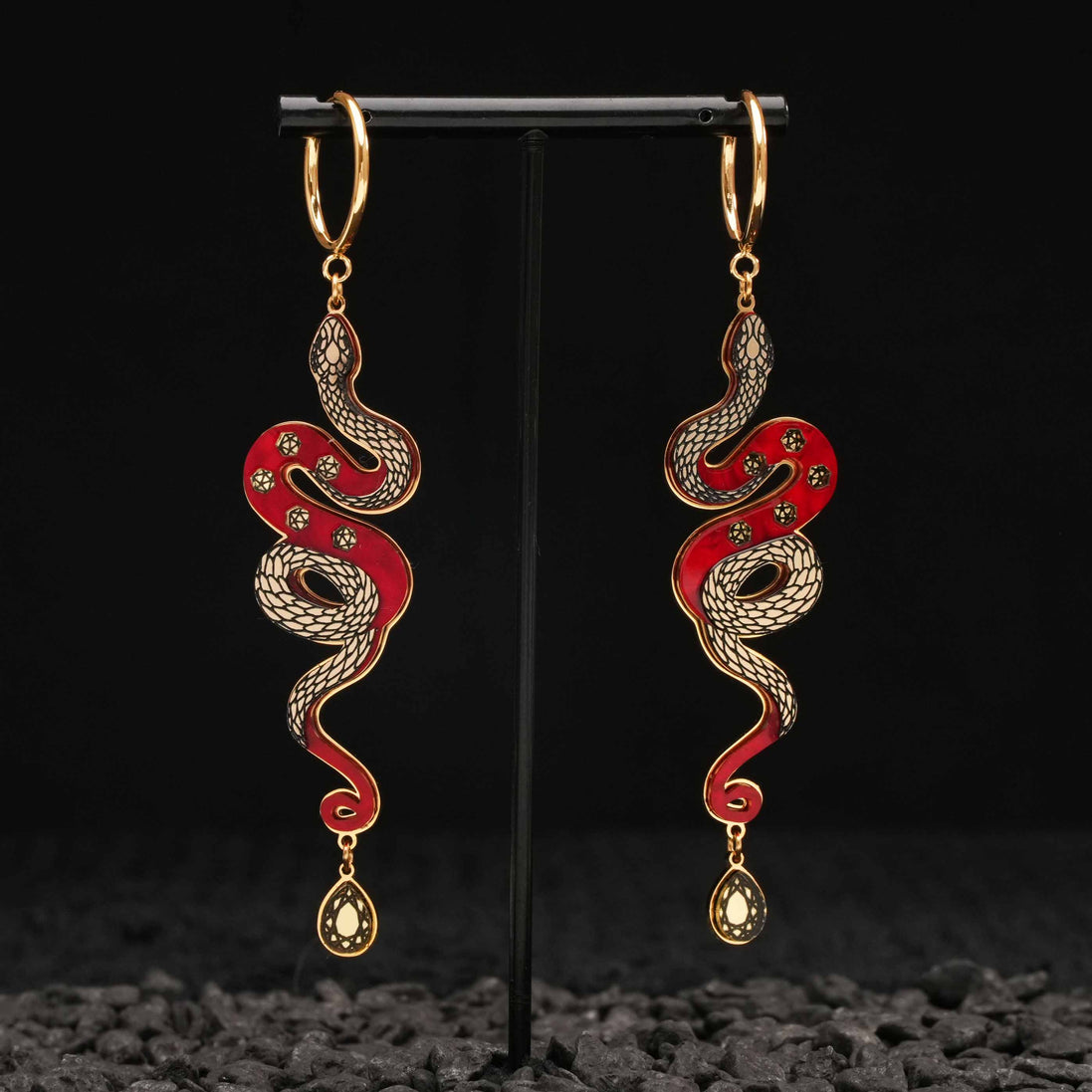 Red and gold Snake-shaped earrings on a black stand with a dark textured background