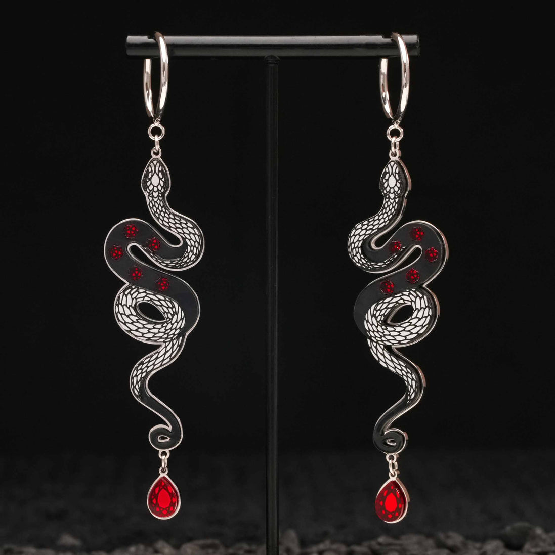 Pair of silver snake-shaped black earrings with red gemstones on a black stand.