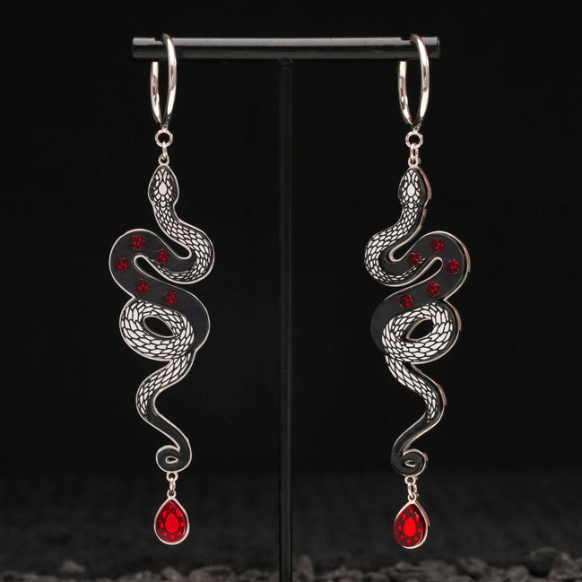 Pair of silver snake-shaped black earrings with red gemstones on a black stand.