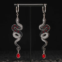 Load image into Gallery viewer, Pair of silver snake-shaped black earrings with red gemstones on a black stand.