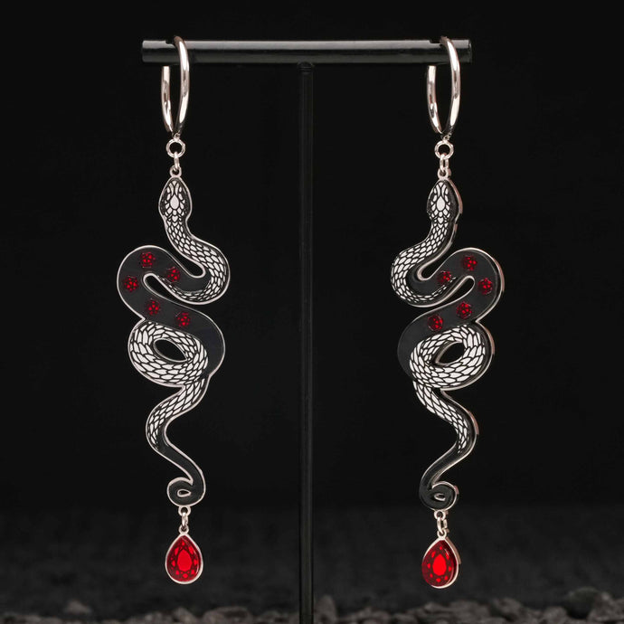 Pair of silver snake-shaped black earrings with red gemstones on a black stand.
