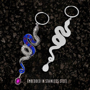 Two snake-shaped silver hoop earrings, one blue and one white, on a dark textured background.