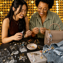 Load image into Gallery viewer, Two women smiling and crafting jewelry together at a table, surrounded by beads, charms, and trays of findings. A taupe handbag with a beaded hanging charm and a denim jacket are visible on the table, set against a gold sequin wall backdrop.
